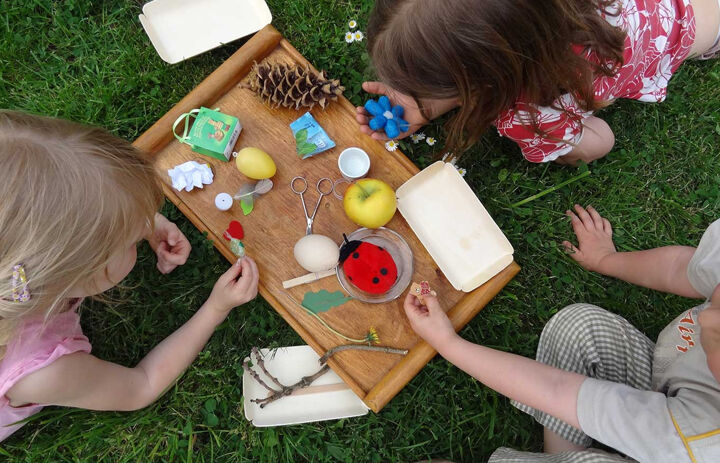 Drei Kinder liegen auf dem Bauch um ein Holbrezbrett herum auf einer Wiese. Auf dem Brett liegen verschiedene Gegenstände, wie eine Schere, ein Apfel, ein Plüschtier-Marienkäfer und ein Tannenzapfen.