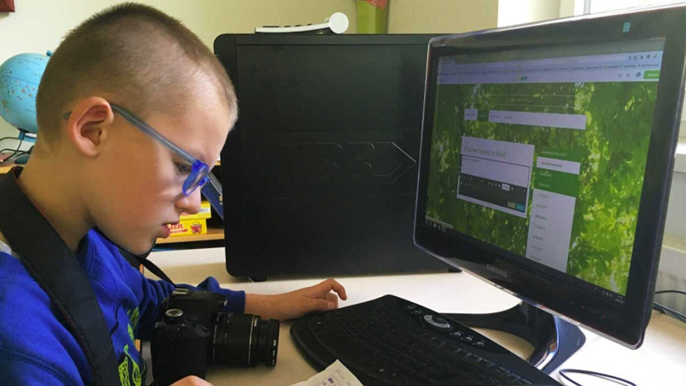 Ein Junge mit blauer Brille sitzt vor einem PC. Vor ihm auf dem Tisch liegt eine Kamera und er schaut konzentriert auf ein Blatt Papier mit Notizen. Der PC-Bildschirm ist angeschalten.