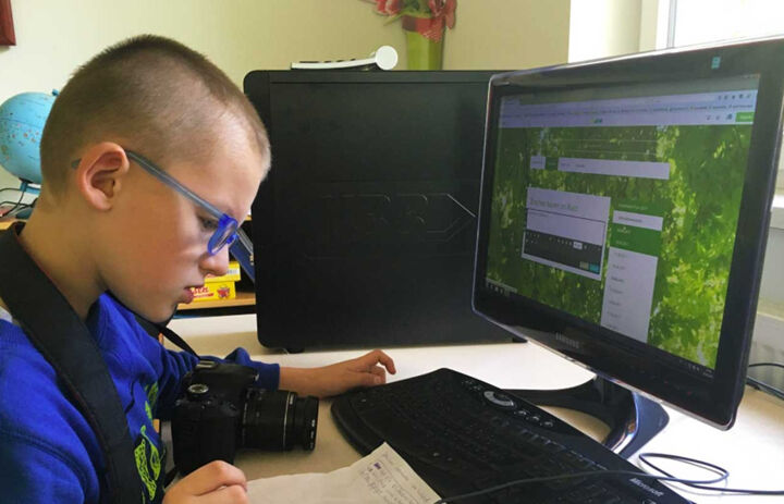 Ein Junge mit blauer Brille sitzt vor einem PC. Vor ihm auf dem Tisch liegt eine Kamera und er schaut konzentriert auf ein Blatt Papier mit Notizen. Der PC-Bildschirm ist angeschalten.