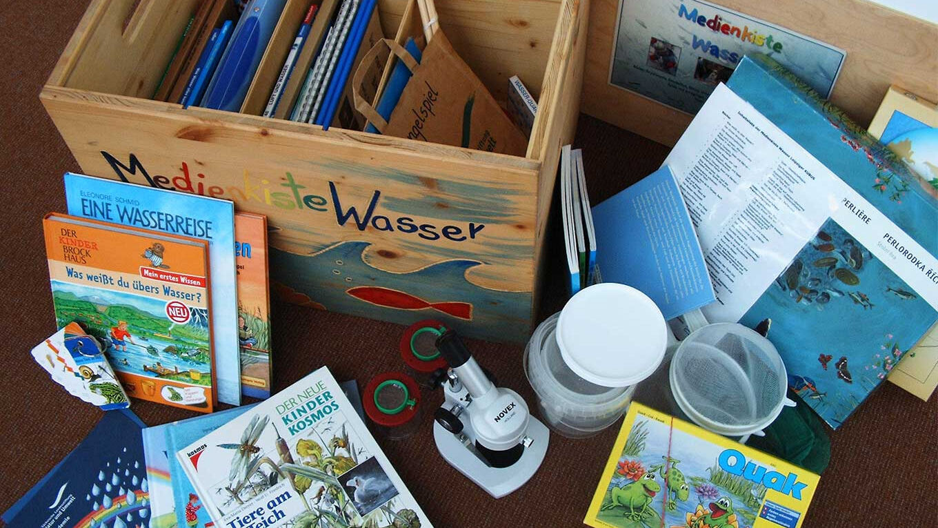 Eine Holzkiste mit der bunten Aufschrift "Medienkiste Wasser"steht auf dem Boden. Um sie herum sind informative Bücher zum Thema Wasser, ein kleiner Käscher und ein Kindermikroskop verteilt.