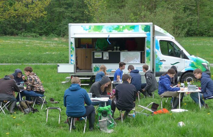 Schüler:innen sitzen in Gruppen an Tischen vor dem Umweltmobil.