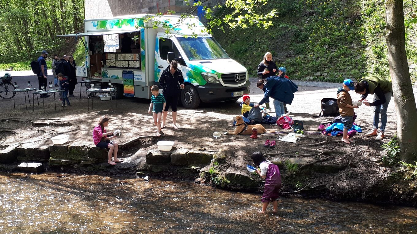 Das Umweltmobil steht am Ufer der Prießnitz. Erwachsene und Kinder sitzen am Ufer oder keschern im Wasser.