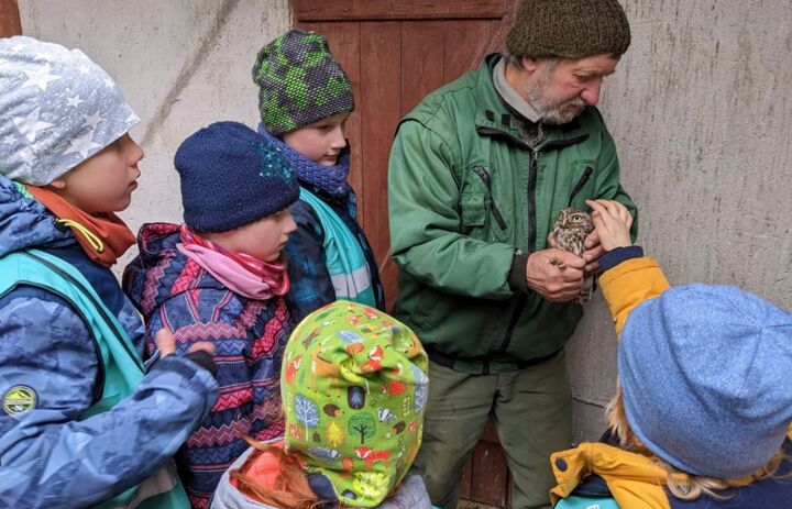 Mitarbeiter des Naturschutzzentrums hält einen Steinkauz in den Händen, eine Gruppe Kinder sieht aufmerksam zu.