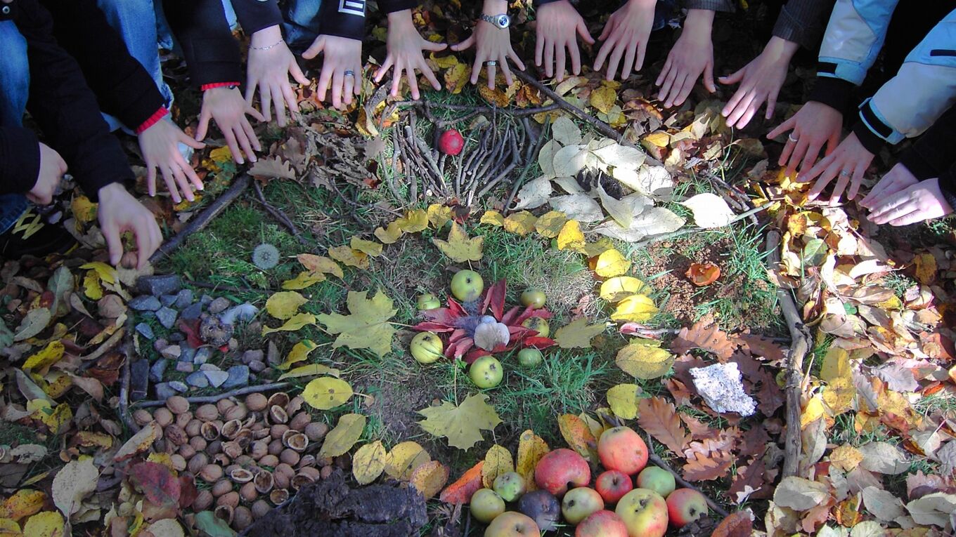 Ein Landartbild aus Naturmaterial wie Äpfel, Nüsse, Zweige, Blätter. Diese ergeben einen Kreis, der zum Teil von Händen der Teilnehmenden berührt wird.