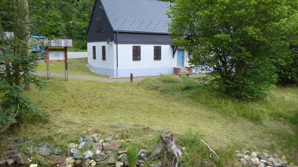Foto der Flussperlmuschelzuchtstation. Ein weißes Gebäude mit schwarzem Dach in der Größe eines Einfamilienhauses. Im Vordergrund ein Bach.