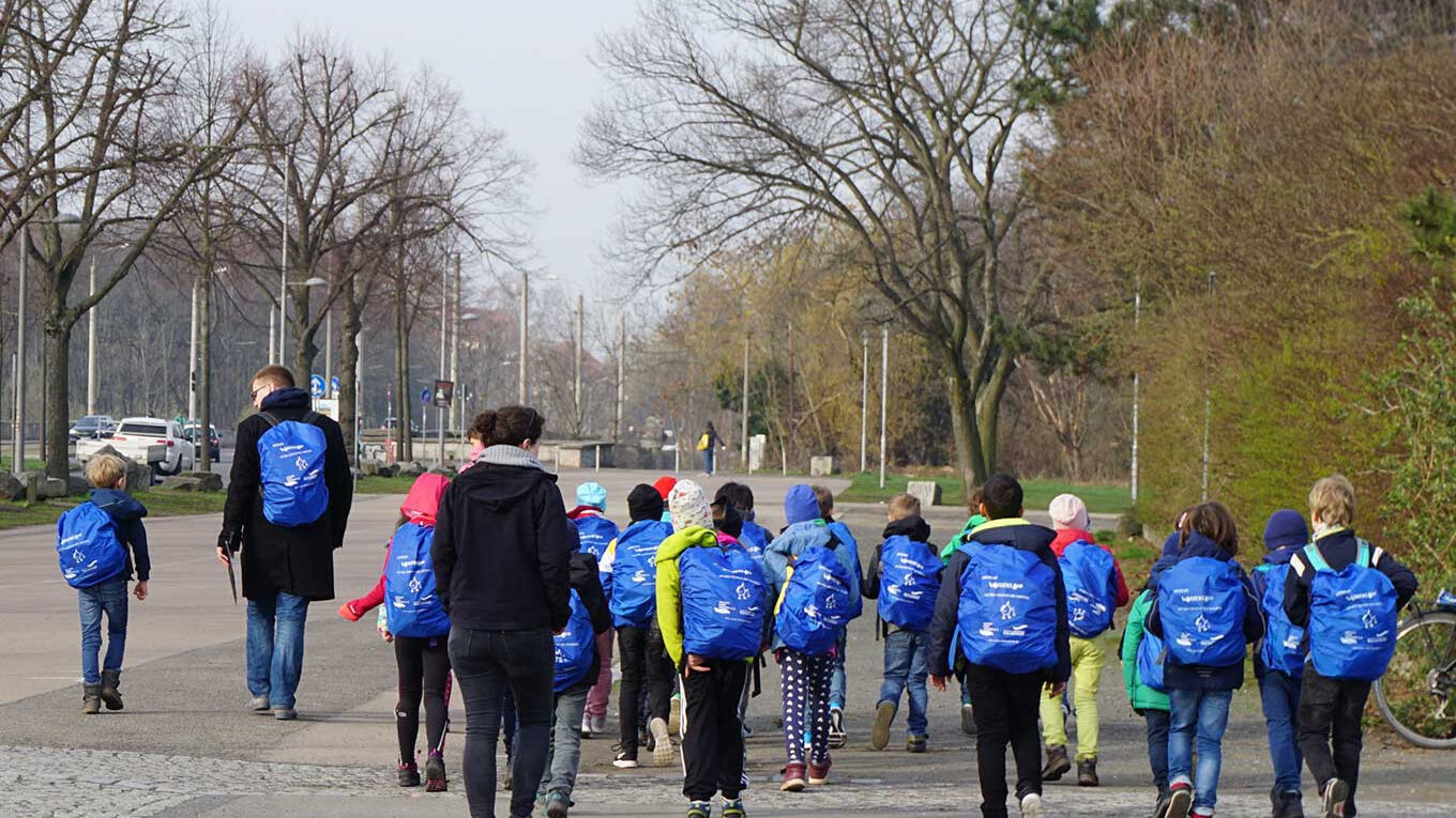 Einge Gruppe von Kindern läuft auf dem Gehsteig. Sie alle tragen den gleichen blauen Rucksack. Die Gruppe wird von zwei Erwachsenen begleitet.