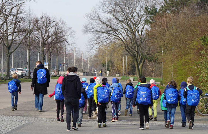 Einge Gruppe von Kindern läuft auf dem Gehsteig. Sie alle tragen den gleichen blauen Rucksack. Die Gruppe wird von zwei Erwachsenen begleitet.