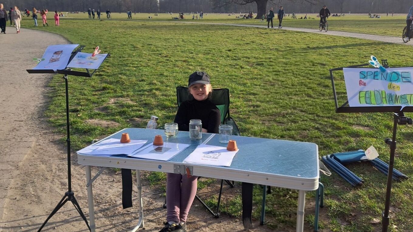 Anca sitzt an einem Klapptisch mit selbst gemalten Infomaterialien und Plakaten  in einem Park (Hinweis: Im Foto ist  eine unbeteiligte Person mit Hilfe von KI entfernt worden).