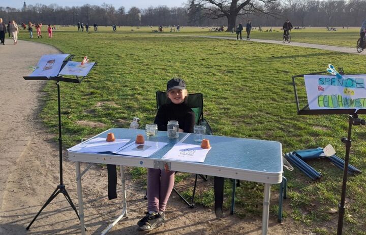Anca sitzt an einem Klapptisch mit selbst gemalten Infomaterialien und Plakaten  in einem Park (Hinweis: Im Foto ist  eine unbeteiligte Person mit Hilfe von KI entfernt worden). - Link: Großes Engagement für den Feldhamster