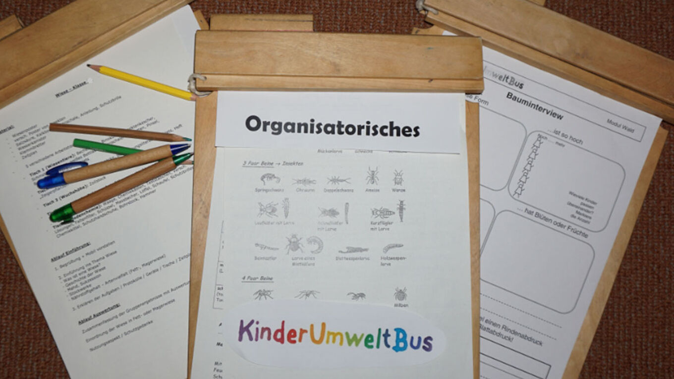 Ein Holklemmbrett mit der Überschrift "Organisatorisches" und der bunten Aufschrift "KinderUmweltBus" liegt auf zwei anderen Klemmbrettern. Auf dem linken Klemmbrett liegen fünf Stifte verstreut.