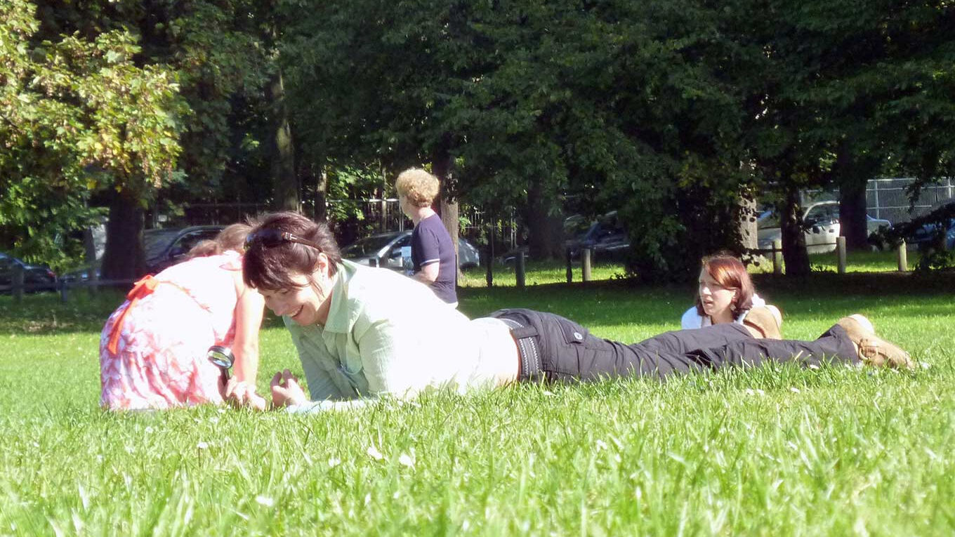 Eine Frau liegt im Gras und lacht. Hinter ihr sieht man mehrere Erwachsene auf der Wiese liegen.