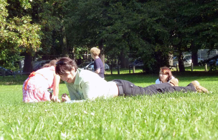 Eine Frau liegt im Gras und lacht. Hinter ihr sieht man mehrere Erwachsene auf der Wiese liegen. - Link: Mit Grundschüler:innen auf der Wiese