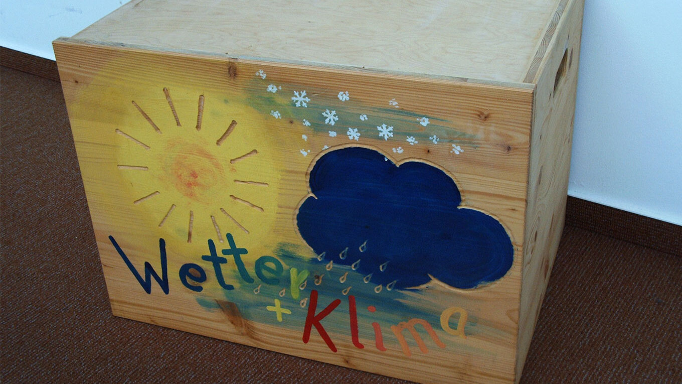 Eine Holzkiste steht auf dem Boden. Auf ihrer Voderseite ist eine gelbe Sonne, dunkelblau Wolke, Regen und Schnee gemalt. Darunter steht in bunten Buchstaben "Wetter + Klima".
