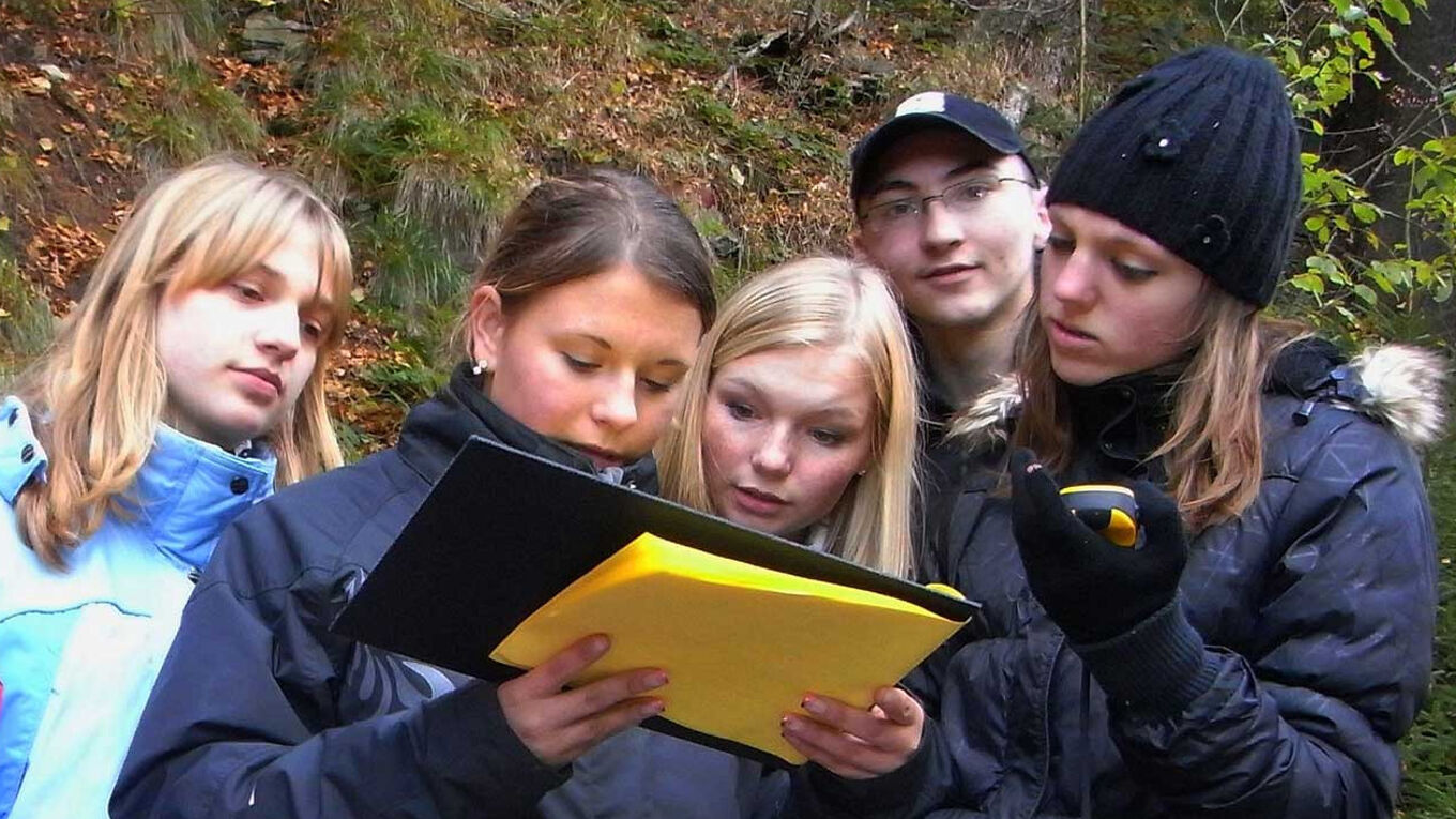 Eine Gruppe von fünf Jugendliche - vier Mädchen und ein Junge, der in die Kamera blickt - konzentrieren sich auf Klemmbrett. Sie stehen im Wald.