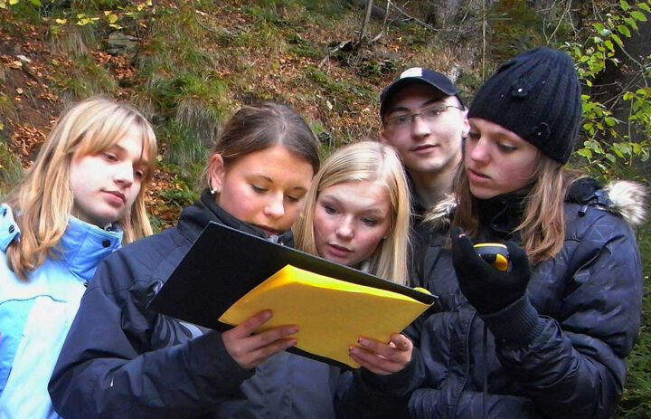 Eine Gruppe von fünf Jugendliche - vier Mädchen und ein Junge, der in die Kamera blickt - konzentrieren sich auf Klemmbrett. Sie stehen im Wald. - Link: Nutzung GPS-gestützer Methoden