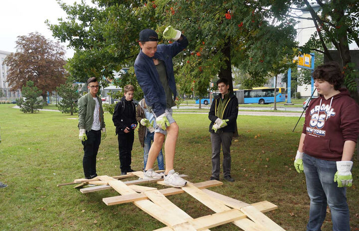 Ein Junge balanciert über eine gewölbte Holzkonstruktion, die auf einer Wiesse liegt. Ihm schaut eine Gruppe anderer Jungen zu.