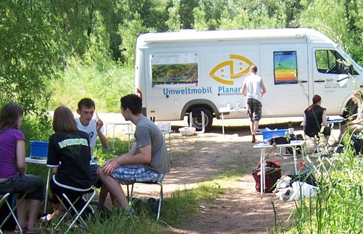 Eine Gruppe Jugendlicher sitz um einen Klapptisch auf einer Wiese. Im Hintergrund steht ein weißer Transport mit der Aufschrift "Umweltmobil Planaria".