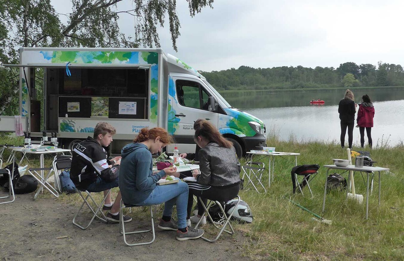 Eine Gruppe von Jugendlichen sitzt um einen Tisch im Freien. Hinter ihnen steht das aufgeklappte Umwelmobil - ein großer weißer Bus. Im Hintergrund ist ein See, an dem zwei Schülerinnen stehen.