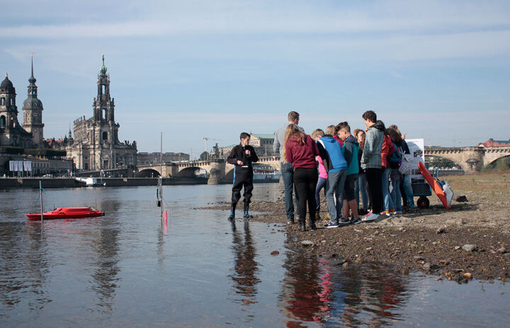 Eine Gruppe Jugendlicher steht am Rand eines Fluss. Ein Mann mit wasserfester Hose und Gummistiefeln steht vor ihnen am seichten Rand des Wassers. Im Hintergrund sieht man eine Brücke und Kirche.