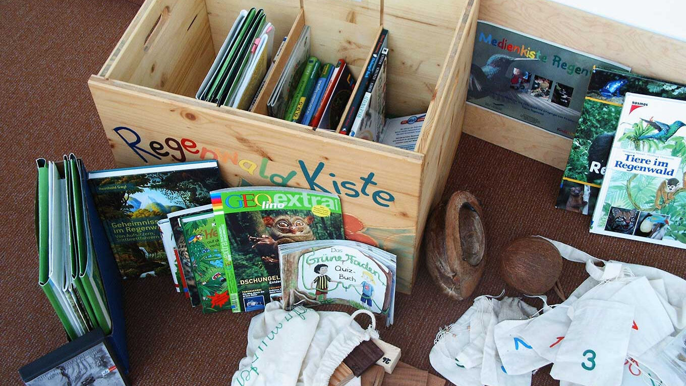Eine Holzkiste mit der Aufschrift "Regenwald Kiste" steht auf dem Boden. Um sie herum liegen Bücher und Zeitschriften zum Thema Regenwald, eine Kokosnuss und weiße Stoffsäckchen mit bunten Nummern.