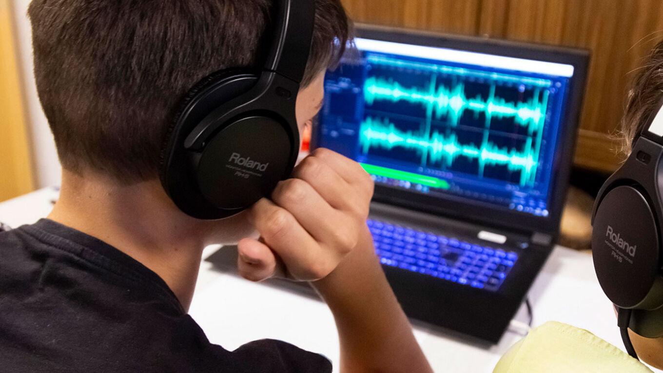 Ein Junge mit großen Kopfhörern sitzt vor einem Laptop. Auf dem Laptop-Bildschirm sind zwei Audiowellenen in einem Bearbeitungsprogramm zu sehen.