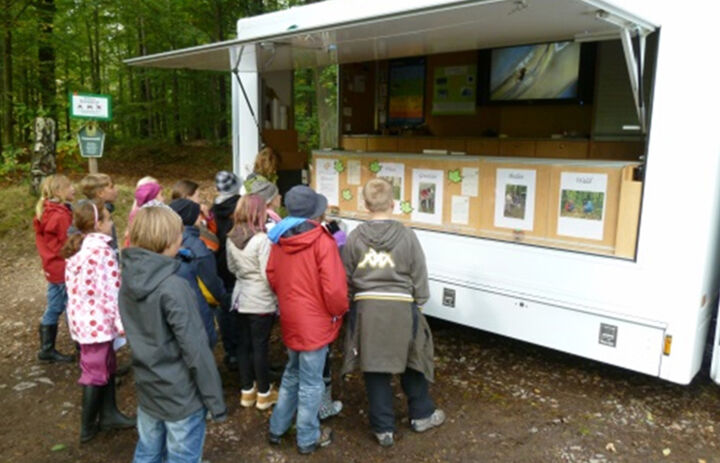 Eine Gruppe Kinder steht vor einem aufgeklapptem Transporter und betrachten aufgehängte Info-Blätter. Der Transporter steht im Wald.