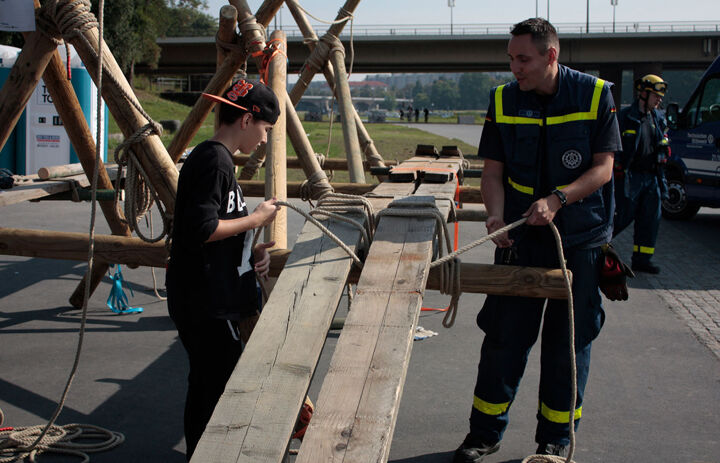 Ein Junge hält ein Seil in der Hand, dass er um ein Brett einer großen Holzkonstruktion wickelt. Ihm gegenüber steht ein Feuerwehrmann in Uniform, der das andere Ende des Seils hält.