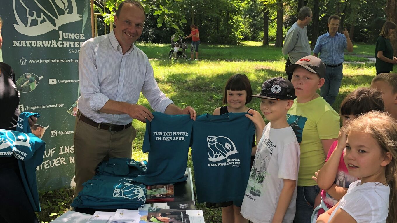 Zu sehen ist der sächsische Umweltminister Wolfram Günther, der einer Gruppe Kindern aus dem Programm Junge Naturwächter Aktions-Shirts mit dem Logo des JuNA-Programms überreicht.
