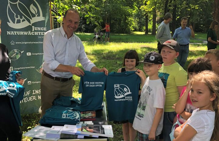 Zu sehen ist der sächsische Umweltminister Wolfram Günther, der einer Gruppe Kindern aus dem Programm Junge Naturwächter Aktions-Shirts mit dem Logo des JuNA-Programms überreicht. - Link: Junge Naturwächter Sachsen